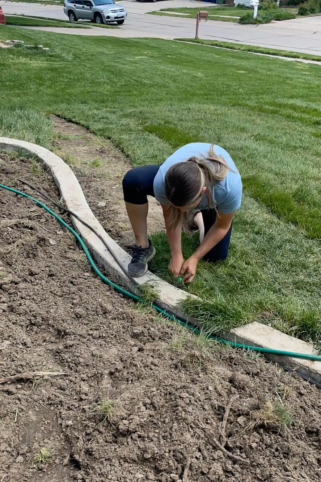 Woman kneeling in the grass while working along a freshly poured concrete garden edge. She’s securing a green garden hose or drip irrigation tubing, prepping the border between lawn and flower bed.